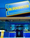 Blockbuster store exterior with blue facade, yellow ticket logo, and glass entrance under a clear sky.