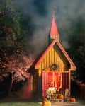 Small wooden chapel with red roof, surrounded by trees and fog, lit pumpkins on porch.