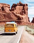 Yellow van with surfboards on roof driving through desert canyon road with red rock formations.
