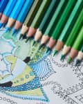 Colored pencils in shades of blue and green on a partially colored intricate mandala design.