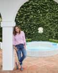Woman in a pink shirt and jeans leaning against a white archway near a small round pool and lush greenery.