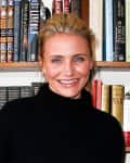 A person smiling in front of a bookshelf filled with various books, wearing a black turtleneck sweater.