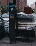 Furniture and mattresses wrapped in plastic stacked on a city sidewalk near parked cars and a parking meter.