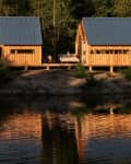 Two wooden cabins with metal roofs by a lake, surrounded by trees, with a person relaxing on a deck.