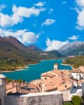Italian village with stone buildings, a tower, and a flag overlooking a turquoise lake and mountains under a blue sky.