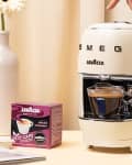 Smeg espresso machine with a cup of coffee, Lavazza pods, and a vase with flowers on a beige table.