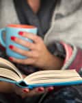Person wrapped in a blanket holding a blue mug, reading a book.