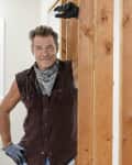 Man in a sleeveless shirt and bandana leaning on wooden beams in a bright room.
