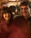 Two people smiling in a crowded bar, with warm lighting and blurred background.