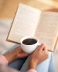 Person holding a book and a cup of coffee, with a croissant on a table in the background.