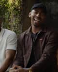Two men sitting outdoors, one in a white shirt and glasses, the other in a dark jacket and cap, smiling near a fire pit.