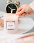 Hand lighting a "Hotel Lobby" candle on a white table with books, petals, and an alarm clock.