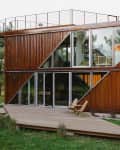 Modern two-story container home with large glass windows, wooden deck, and outdoor chairs surrounded by greenery.