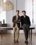 Two men standing in a bright room with wooden floors, wicker pendant lights, and a dark wooden table.