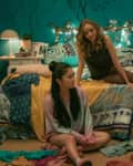 Three girls in a cozy, teal bedroom with fairy lights, sitting and talking on a bed and chair, surrounded by colorful decor.