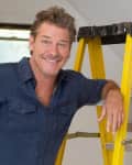 Man in a blue shirt smiling, leaning on a yellow ladder indoors.