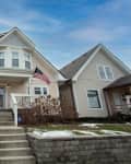 Two-story beige house with front porch, American flag, and stone steps, flanked by similar homes.