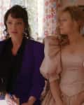Two women in 80s-style dresses sit on a bed in a floral wallpapered room.