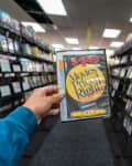 Hand holding a Blockbuster DVD case in a video rental store aisle filled with movie cases.