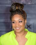Woman in a bright green top with braided updo and hoop earrings, smiling against a gray brick wall.