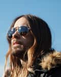 Person with long hair and sunglasses wearing a fur-lined jacket, looking up at a clear blue sky.