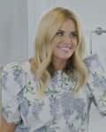 Woman with blonde hair in floral dress smiling in a bathroom with a towel rack and soap dispenser.