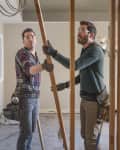Two men in a partially renovated room holding a wooden beam, wearing tool belts and casual clothing.