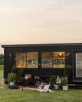 Modern black tiny house with large windows, string lights, and outdoor seating on a grassy field at sunset.