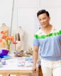 Man in a blue and green shirt standing by a table with colorful decor items, flowers, and mannequins in a bright studio.