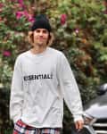 Person in a white "ESSENTIALS" sweatshirt and plaid pants, standing outdoors near a car and flowering bushes.