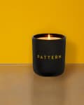 Black candle with "PATTERN" text, lit on a reflective surface against a yellow background.