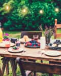 Outdoor dining table with assorted berries, pastries, drinks, and flowers, set against a backdrop of greenery and string lights.