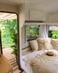 Cozy camper interior with a bed, fluffy pillows, straw hat, and a view of lush greenery through the open door.