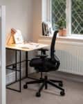 Home office under stairs with desk, black chair, lamp, and potted plant by window.
