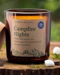 Brown glass candle labeled "Campfire Nights" with cork lid, surrounded by small white stones and moss on a wooden surface.