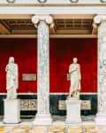 Three marble statues stand between ornate columns against a red wall in a classical gallery setting.