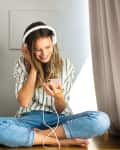 Woman in striped shirt and jeans sitting cross-legged, listening to music on headphones, surrounded by plants.