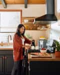 Woman in a cozy kitchen pouring coffee, with fruit and plants on wooden countertops.