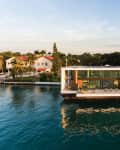 Modern floating house on water with large windows, docked boat, and lush greenery in the background.