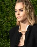 Person with wavy blonde hair wearing a black textured jacket and layered necklaces, standing in front of green foliage.