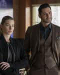Two people standing in a room with wooden furniture and blinds, one in a black jacket and the other in a brown suit.