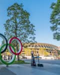 Olympic rings sculpture in a park with trees and a stadium in the background.