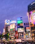 Busy urban scene in Shibuya, Tokyo, featuring bright billboards, shops, and a crowd of pedestrians at dusk.