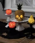 Fruits carved as jack-o'-lanterns on cake stands, including pineapple, apple, and pear, on a wooden table with black lace.
