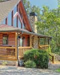 Log cabin with large triangular windows, stone chimney, and wooden porch surrounded by trees and greenery.
