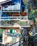 Two-story house made of glass bottles and wood, surrounded by trees and a stone pathway.