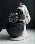 Stand mixer with a wire whisk attachment and textured black mixing bowl on a marble countertop.