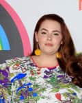 Person with long red hair wearing a floral print top and yellow pom-pom earrings, standing in front of a colorful backdrop.