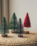 Four miniature Christmas trees, three green and one red, on a woven surface near a window.