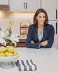 Woman in a kitchen with white cabinets, vase of lilies, bowl of lemons, and a striped towel on the counter.